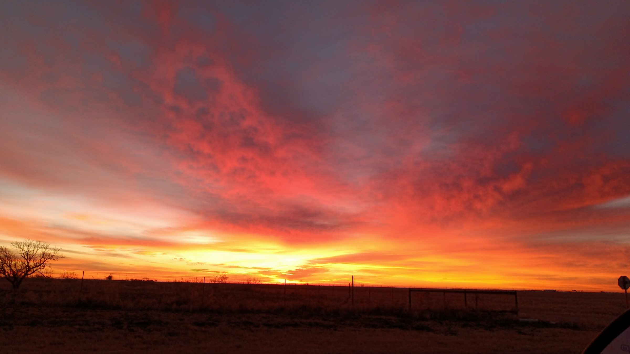 Fiery red and orange clouds fill the sky above flat ranch land and a fence line at sunset