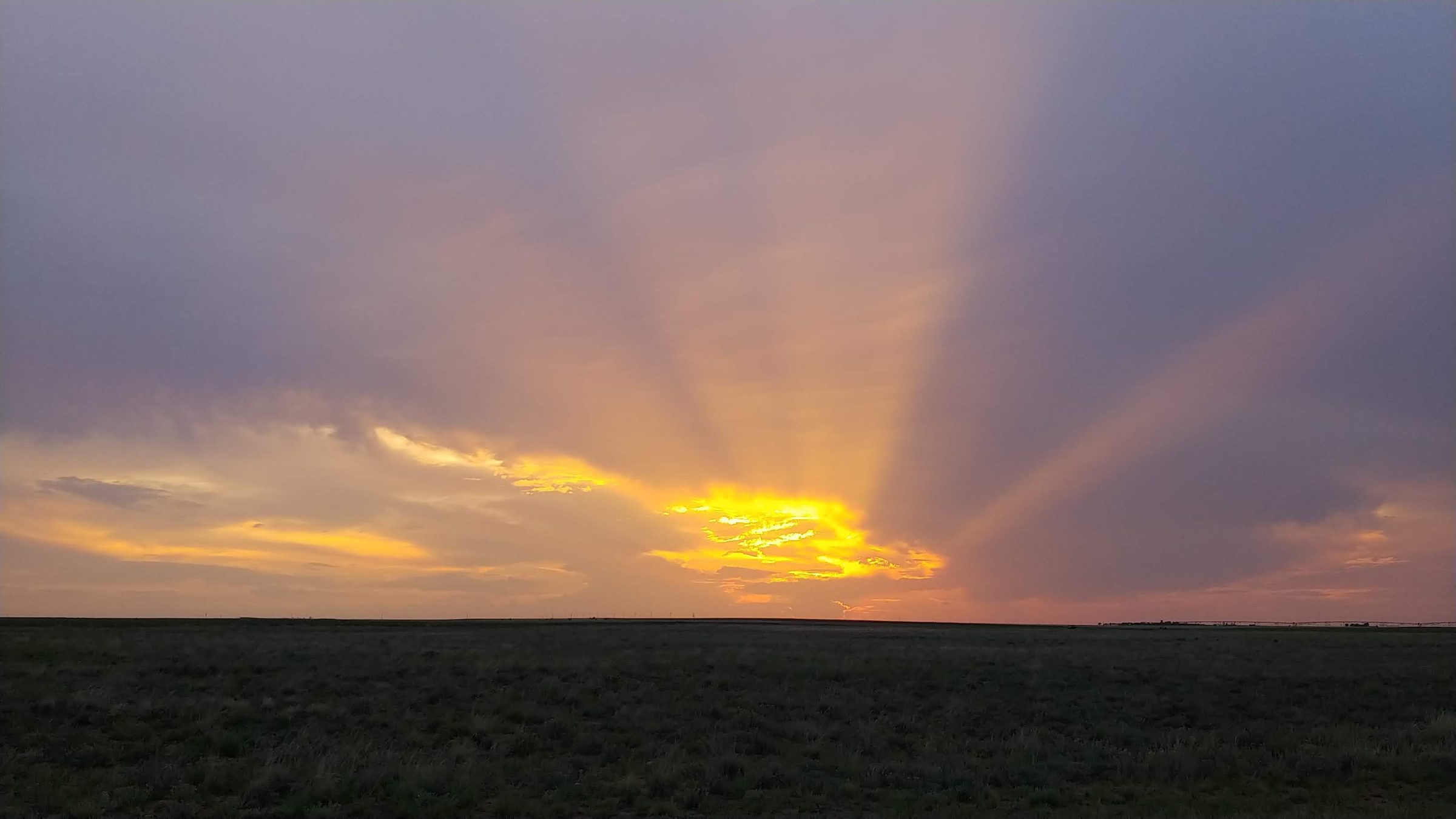 Golden sunbeams fan out from behind gray clouds over a wide, flat field