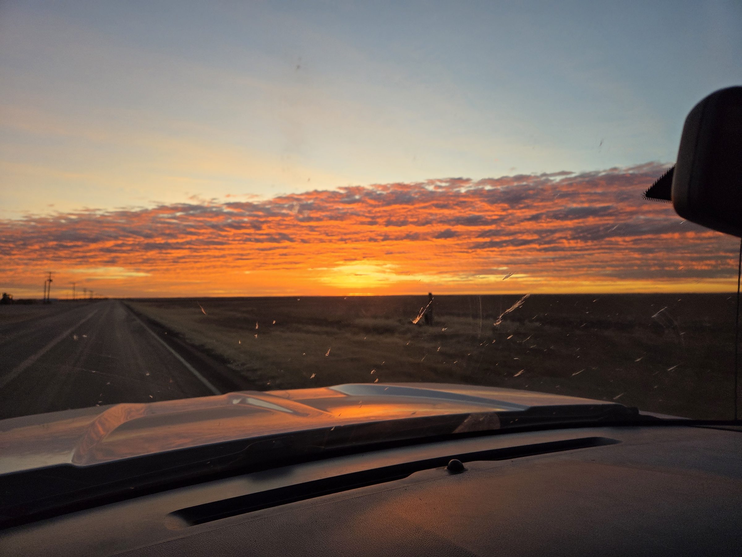 Orange sun on the horizon under a band of glowing clouds, seen over the hood of a truck on a straight two-lane highway